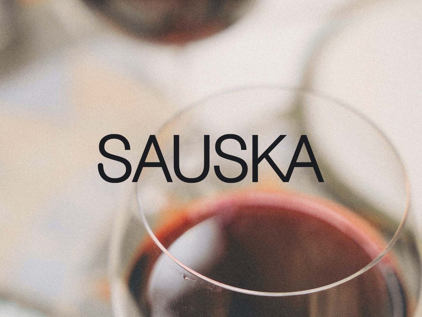 Nahaufnahme eines Rotweinglases mit dem Schriftzug »SAUSKA« vor hellem, unscharfem Hintergrund.