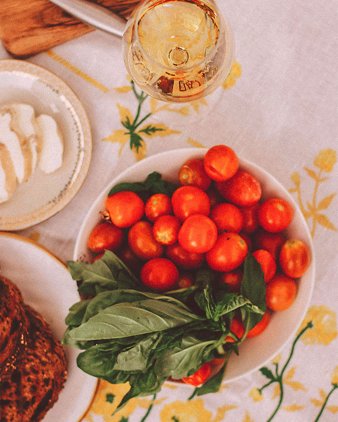 Schale mit frischen Kirschtomaten und Basilikum neben einem Glas Weißwein und Mozzarella auf einer sommerlich gedeckten Tafel.