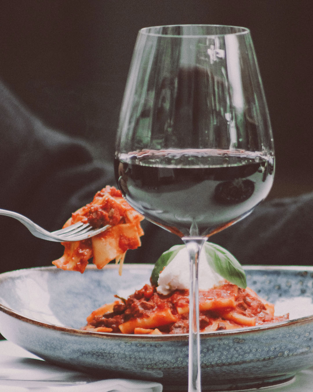 Glas Rotwein neben einem Teller Pasta mit kräftiger Tomatensauce, Mozzarella und frischem Basilikum.